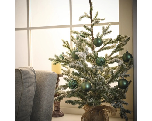 Decoratieve kerstboom met groene ballen en jutezak op een tafel voor een raam.