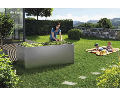 Tuinscene met een metalen verhoogde tuinbak beplant met groenten en een vrouw die groenten oogst. Twee kinderen spelen op een deken in het gras.