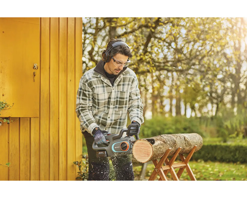 Man zaagt een stuk hout op een zaagbok met een accukettingzaag, terwijl hij een veiligheidsbril en hoofdtelefoon draagt.
