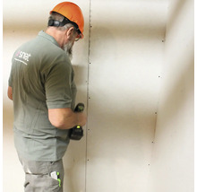 Een man met een veiligheidshelm monteert een gipsplaatwand met een accuschroevendraaier.