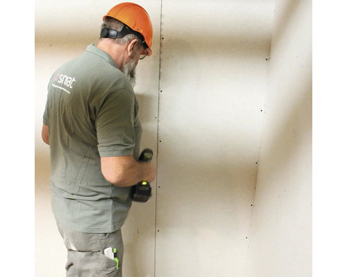 Een man met een veiligheidshelm monteert een gipsplaatwand met een accuschroevendraaier.