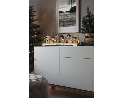 Kerstscène met een verlicht houten dorp op een dressoir, naast een kerstboom en een ingelijste foto.