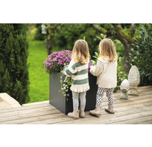 Twee kinderen staan naast een grote, donkere plantenbak met violette bloemen op een houten terras.