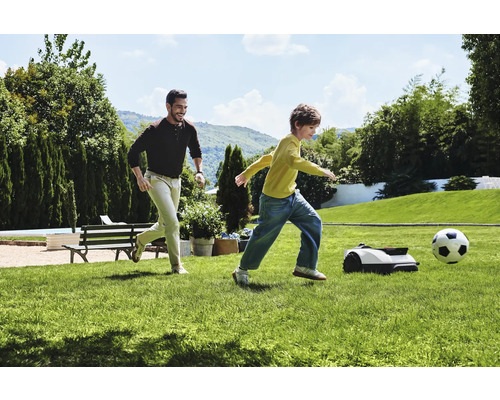 Man en kind spelen voetbal met robotmaaier in de tuin.