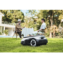 Scene met een robotmaaier in de tuin, terwijl twee mensen aan een tafel zitten