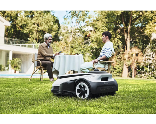 Scene met een robotmaaier in de tuin, terwijl twee mensen aan een tafel zitten