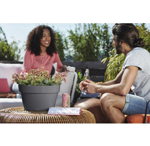 Buitenscène met een grijze plantenpot op een tafel met planten en twee mensen op de achtergrond.