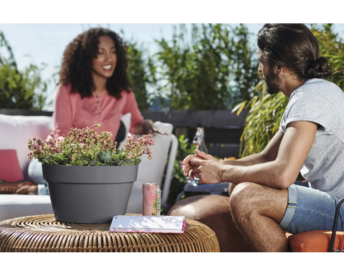 Buitenscène met een grijze plantenpot op een tafel met planten en twee mensen op de achtergrond.