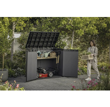 Tuinscene met een vrouw die een kist met planten draagt naast een gereedschapskast gevuld met tuingereedschap en een grasmaaier