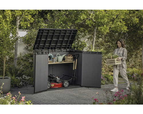 Tuinscene met een vrouw die een kist met planten draagt naast een gereedschapskast gevuld met tuingereedschap en een grasmaaier