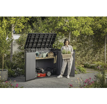 Tuinscene met tuinhuis, tuingereedschap en een vrouw die een kist met planten vasthoudt.