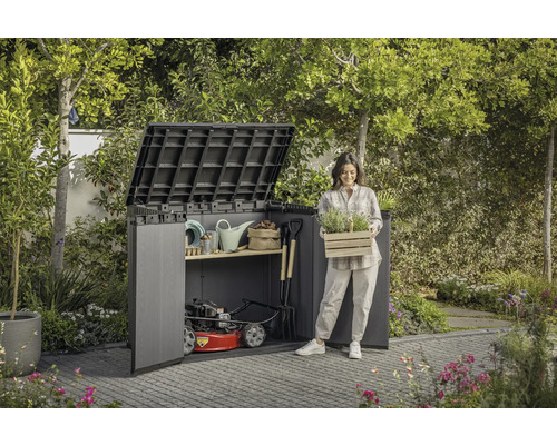Tuinscene met tuinhuis, tuingereedschap en een vrouw die een kist met planten vasthoudt.