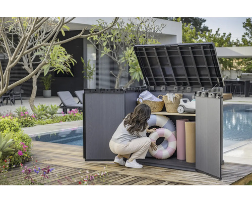 Tuinscene met een vrouw die een kunststof tuinkast met twee planken opent, waarin zwembanden, handdoeken en manden staan.