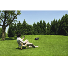 Een man zit in de tuin op een stoel en bedient een gazonrobot met zijn smartphone.