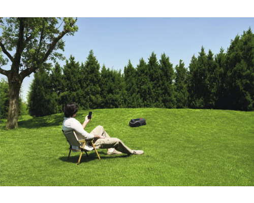 Een man zit in de tuin op een stoel en bedient een gazonrobot met zijn smartphone.