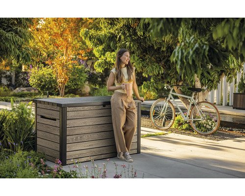 Tuinscene met een vrouw die tegen een houten opbergbox leunt en een koffiekop vasthoudt.