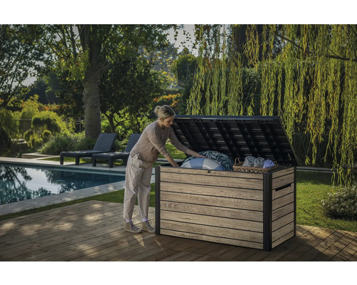 Een vrouw opent een tuinkist op een houten terras naast een zwembad en een tuin.