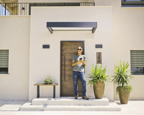 Moderne entree met een voordeur in houtlook, een donkere luifel en buitenwandlampen. Een man staat voor de deur en gebruikt zijn smartphone.