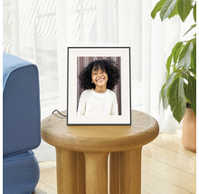 Digitale fotolijst op een ronde houten bijtafel in een lichte woonkamer naast een kamerplant.