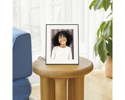 Digitale fotolijst op een ronde houten bijtafel in een lichte woonkamer naast een kamerplant.