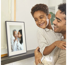Een vader houdt zijn glimlachende zoon vast, beiden kijken naar een digitale fotolijst op een plank met een familiefoto.
