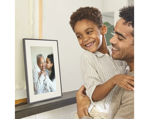 Een vader houdt zijn glimlachende zoon vast, beiden kijken naar een digitale fotolijst op een plank met een familiefoto.