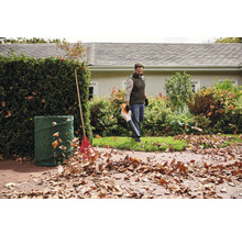 Een man gebruikt een bladblazer in de tuin, naast een tuinafvalzak en een hark.