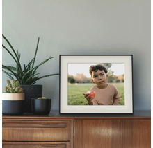 Zwarte fotolijst met passe-partout op een houten dressoir naast verschillende groene kamerplanten tegen een grijze muur.