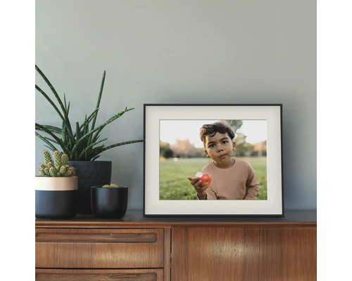 Zwarte fotolijst met passe-partout op een houten dressoir naast verschillende groene kamerplanten tegen een grijze muur.