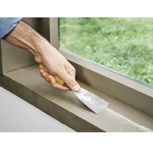 Een hand met een plamuurmes dicht een vensterbank af.