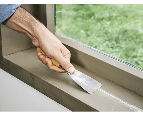 Een hand met een plamuurmes dicht een vensterbank af.