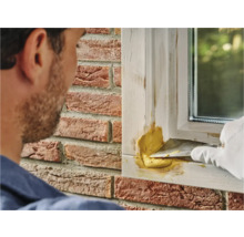 Ambachtsman plamuurt raamkozijn met vulmiddel, met op de achtergrond een bakstenen huis.