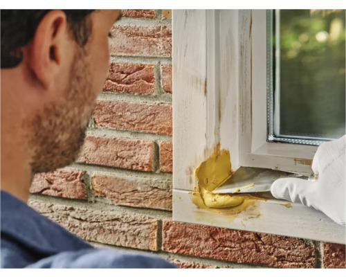 Ambachtsman plamuurt raamkozijn met vulmiddel, met op de achtergrond een bakstenen huis.