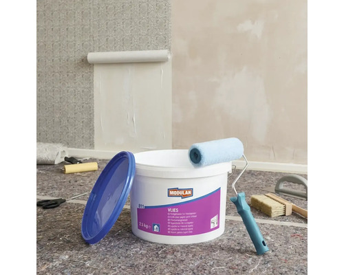 Scene met een emmer vliesbehanglijm, een verfroller, een behangborstel en een begonnen behang aan de muur