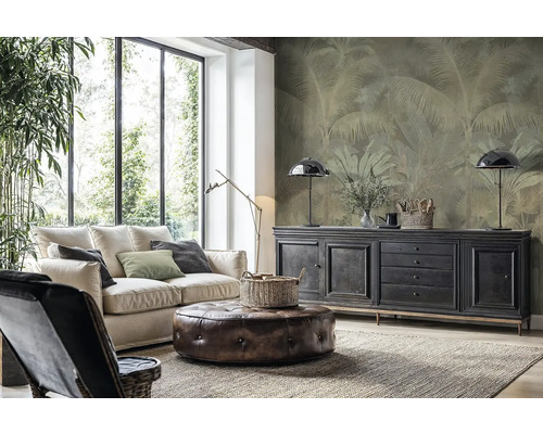 Moderne woonkamer met lichte bank, leren poef en vliesbehang met palmenmotief op de wand achter een dressoir.