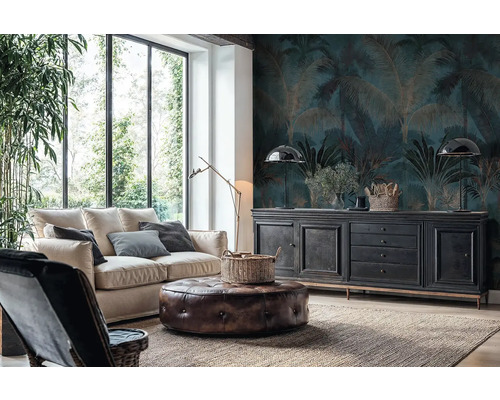 Woonkamer met fotobehang met palmenpatroon in donkerblauw en goud, een lichte bank, leren poef en zwart dressoir voor grote ramen.