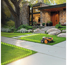 Een robotmaaier op een gazon voor een huis met een stenen gevel en een grote boom.