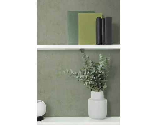 Vliesbehang met fijne structuur in donkergroen op een muur achter een witte wandplank met boeken en een vaas met eucalyptus.