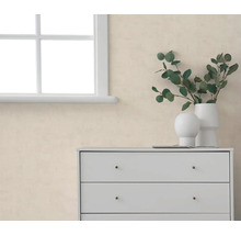 Beige behang met structuur in een lichte kamer naast een wit raam en een witte commode met vazen en eucalyptustakken.