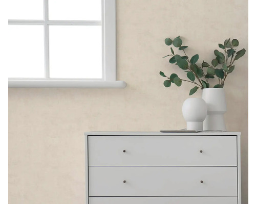 Beige behang met structuur in een lichte kamer naast een wit raam en een witte commode met vazen en eucalyptustakken.