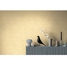 Behang met textuur in warm geel op een muur achter een dressoir met een decoratieve vogelfiguur, vaas met katoentak en kaarsenhouders.