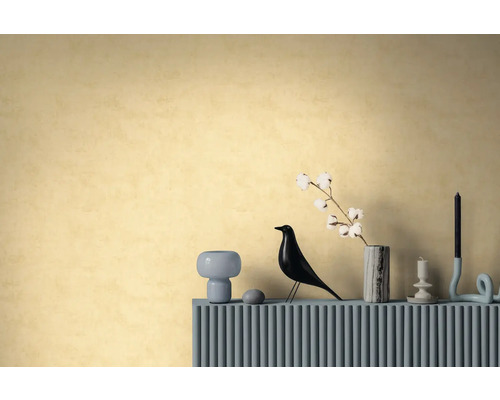 Behang met textuur in warm geel op een muur achter een dressoir met een decoratieve vogelfiguur, vaas met katoentak en kaarsenhouders.