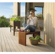 Scene op een terras met een verhoogde tuinbak en een vrouw zittend op een kruk die van een drankje geniet