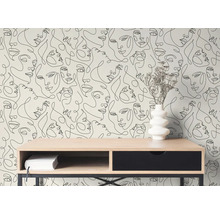 Modern behang met zwarte lijntekeningen van gezichten achter een houten bureau met een witte vaas en droogbloemen in een lichte kamer.