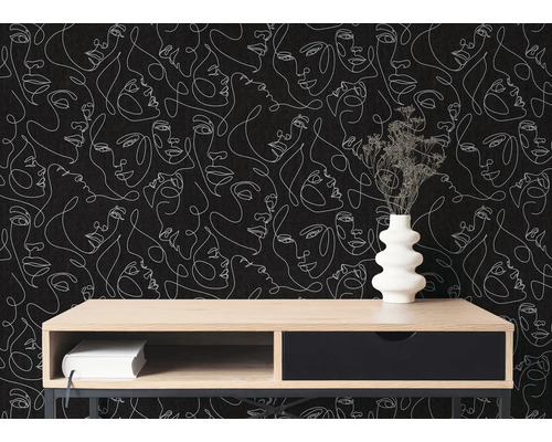 Modern interieur met behang met witte one-line art gezichten op een zwarte achtergrond. Ervoor staat een houten dressoir met een vaas.