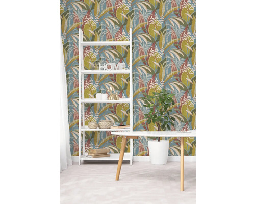 Vliesbehang met botanisch bladpatroon in groen en bruin op blauwe achtergrond, witte stellingkast en houten tafel in een lichte kamer.