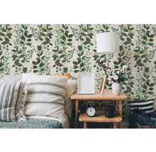 Vliesbehang met groen bladerpatroon in een lichte slaapkamer met houten tafel, lamp en kussens.
