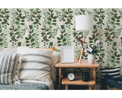 Vliesbehang met groen bladerpatroon in een lichte slaapkamer met houten tafel, lamp en kussens.