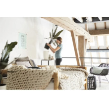 Licht, open appartement met houten balkenplafond en een vrouw die een kamerplant in een sierpot verpot.