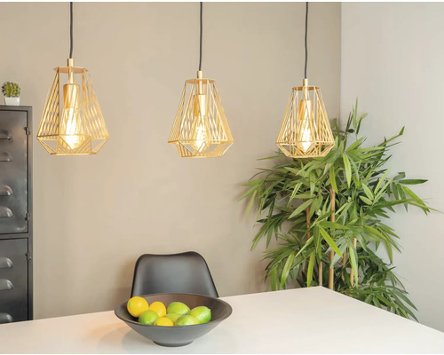 Drie goudkleurige hanglampen boven een eettafel met fruitschaal en een plant op de achtergrond.